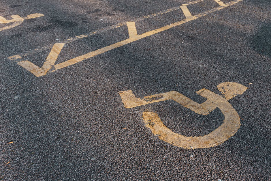 Close-up view of a black asphalt parking surface with yellow painted lines, including a wheelchair symbol indicating accessible parking. The surface appears slightly worn with some scuff marks and dirt, and the yellow paint shows signs of fading and chipping, particularly around the edges of the symbol. The image is taken outdoors under natural lighting, capturing the textured asphalt and clearly visible parking markings. This setting relates to accessibility considerations in parking areas, similar to issues addressed by South Kensington Cleaner in maintaining clean and compliant parking zones, as referenced in the page titled 'Access and parking issues for cleaners in Earls Court Road, SOUTH KENSINGTON Cleaner'.
