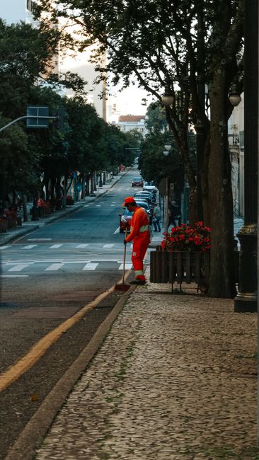 A street scene on Earls Court Road in South Kensington during daytime, showing a worker in a bright red uniform sweeping the cobblestone sidewalk with a long-handled broom. The street features a row of parked cars along the curb, lined with large trees that cast shade over the area. Ornamental street lamps are visible among the trees, and flowers are planted in a small bed adjacent to the sidewalk. The background includes buildings and a partly cloudy sky. The image emphasizes cleanliness and maintenance activities typical of residential or commercial cleaning efforts, with a focus on surface cleaning and tidiness maintained by South Kensington Cleaner, highlighting their role in ensuring hygienic urban environments.