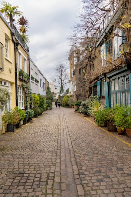 A cobblestone alleyway lined with residential buildings on both sides, featuring a mix of modern and traditional architectural styles. The left side has a cream-colored facade with large windows and balconies adorned with potted plants, while the right side displays a darker, aged brick structure with glass-enclosed extensions. The alley is decorated with various green plants in pots and planters, creating a lush atmosphere. Bare trees with twisted branches are visible overhead, and the sky is overcast with gray clouds, suggesting a calm, quiet residential area. Natural light subtly illuminates the scene, emphasizing the textures of the cobblestones and the greenery. The area appears clean and well-maintained, aligning with professional residential cleaning standards promoted by South Kensington Cleaner.