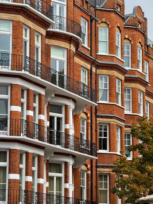 Photograph of a multi-story residential building with red brick exterior walls, featuring white architectural accents around the windows and balconies. The building has multiple levels with black wrought-iron railings on the balconies, some of which are adorned with small potted plants. Large windows with white frames allow natural light into the interiors. A leafy green tree partially obscures the lower floors, adding a touch of nature to the scene. The overall appearance of the building is clean and well-maintained, exemplifying urban residential architecture typical of South Kensington. The image relates to the cleaning services offered by South Kensington Cleaner, highlighting the importance of surface cleaning and maintenance for apartment exteriors or communal areas, consistent with their domestic cleaning and deep cleaning offerings.