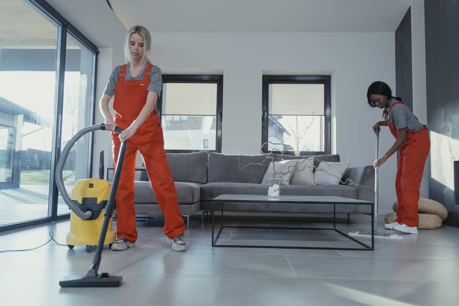 Two professional cleaners from South Kensington Cleaner, dressed in gray shirts and red overalls, performing surface cleaning in a modern living room. The cleaner on the left is using a yellow vacuum cleaner to clean the grey tiled floor, while the cleaner on the right is wiping a glass coffee table with a cloth. The room features a grey fabric sofa with patterned cushions, a black metal frame coffee table, and large windows allowing natural light to illuminate the space. The surfaces appear spotless and well-maintained, with no visible dust or dirt, highlighting a high standard of deep cleaning and sanitisation in a contemporary residential setting.