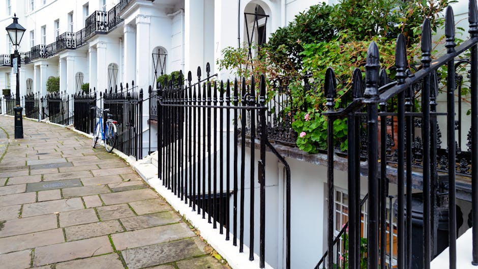 A paved sidewalk made of rectangular stone slabs runs alongside a white residential building in Kensington, featuring black wrought iron railings and planters with green foliage and pink flowers. A bl