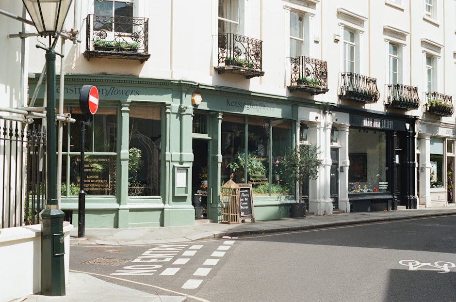 Exterior view of a row of commercial shops on a street in Kensington High Street, featuring large glass windows displaying plants and merchandise, with pastel green and white facades and decorative wrought iron balconies above. The sidewalk is clean, with a blackboard sign near the entrance of the florist shop, and a bicycle lane marked on the road. Natural daylight illuminates the scene, highlighting the well-maintained building surfaces and street furniture, indicative of thorough cleaning and maintenance by South Kensington Cleaner, specializing in surface cleaning, deep cleaning, and sanitisation for residential and commercial spaces.
