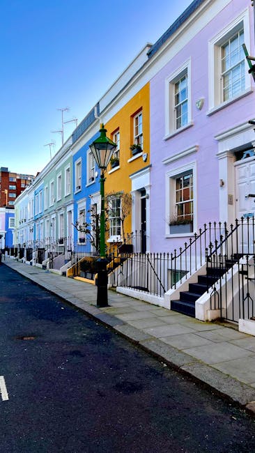 A row of colorful residential terrace houses in Kensington and Chelsea, featuring pastel facades in shades of blue, yellow, lavender, and pink. The buildings have white-framed sash windows, decorative moldings, and black wrought-iron railings along the front steps. A traditional black street lamp stands near the sidewalk, which is made of grey paving stones. The street appears clean and well-maintained, with parked cars visible further down the road. The scene exemplifies the vibrant, historic architecture typical of South Kensington, and reflects the type of environment where professional cleaning, such as that offered by South Kensington Cleaner, may be required for maintaining the appearance and hygiene of residential properties.