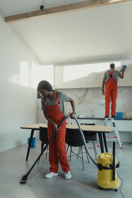 Two professional cleaners from South Kensington Cleaner, dressed in grey t-shirts and orange overalls, are performing cleaning tasks in a modern, well-lit kitchen. One cleaner is using a vacuum cleaner on the light-colored hardwood floor, visibly removing dust and debris. The second cleaner, standing on a small stepladder, is sanitising or wiping down a white, marble backsplash area behind the countertop. The kitchen features a sleek, minimalist design with glossy white cabinets, a wooden ceiling beam, and a dining table with black chairs positioned in the center. Bright natural light flows in through large windows, illuminating the clean, shiny surfaces and creating a fresh atmosphere. This scene exemplifies professional domestic cleaning focused on surface cleaning and hygiene enhancement, aligned with common practices recommended by South Kensington Cleaner to avoid mistakes when booking a Kensington cleaning service.