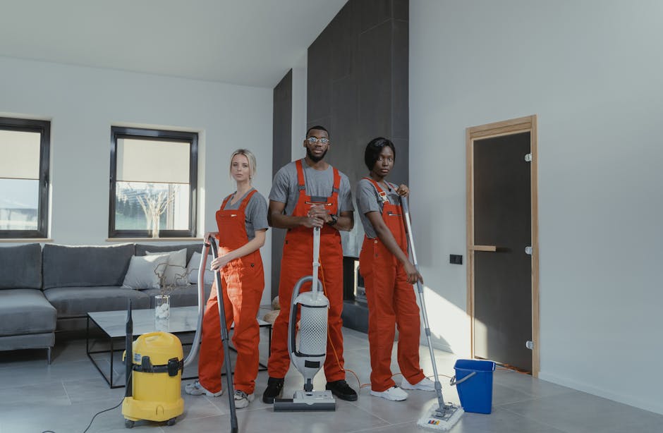 Two professional cleaning staff members in beige uniforms standing indoors against a plain white wall, holding cleaning tools. The woman on the left holds a red and black vacuum cleaner, while the woman on the right has a green-handled mop. Both are smiling and appear ready for domestic or commercial surface cleaning, emphasizing hygiene and efficient cleaning practices in a well-lit, minimalistic environment. Their uniforms feature badges, and the scene highlights a clean, dust-free setting suitable for detailed cleaning or deep sanitisation services offered by South Kensington Cleaner according to their guidelines on booking mistakes for Kensington cleaning services.