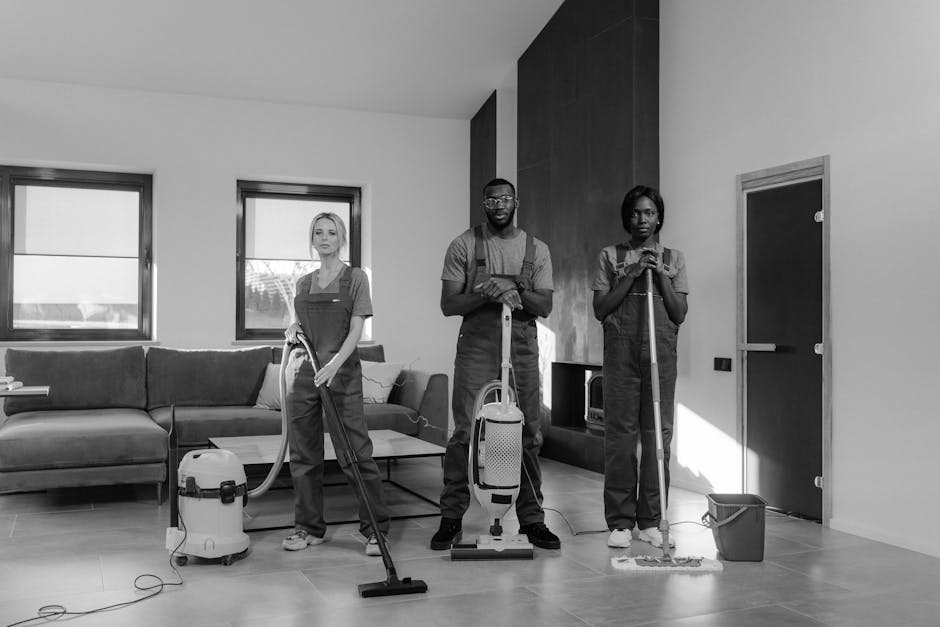Three professional cleaners from South Kensington Cleaner are performing surface cleaning in a modern living room. The room features a light-colored tiled floor that is being vacuumed by the individual in the center, who holds a portable vacuum cleaner. On the left, a female cleaner with blonde hair is standing with a vacuum hose in her hand, near a dark grey fabric sofa placed against a white wall with two large windows letting in natural light. The rightmost cleaner, a woman with dark hair, is using a mop and bucket for floor mopping near a door. Behind them, a sleek accent wall in dark wood panels contrast with the white walls and ceiling, and the room is well-lit with bright natural daylight. Visible cleaning tools include a vacuum cleaner, a mop, and a bucket, all arranged methodically. The scene emphasizes thorough, professional domestic cleaning and surface sanitisation, highlighting attention to hygiene and cleanliness in a contemporary residential setting, with South Kensington Cleaner providing expert cleaning services.